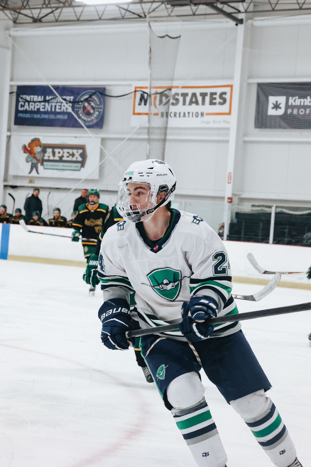 Hockey player in white jersey with teal logo skates on ice during competitive match at indoor rink.