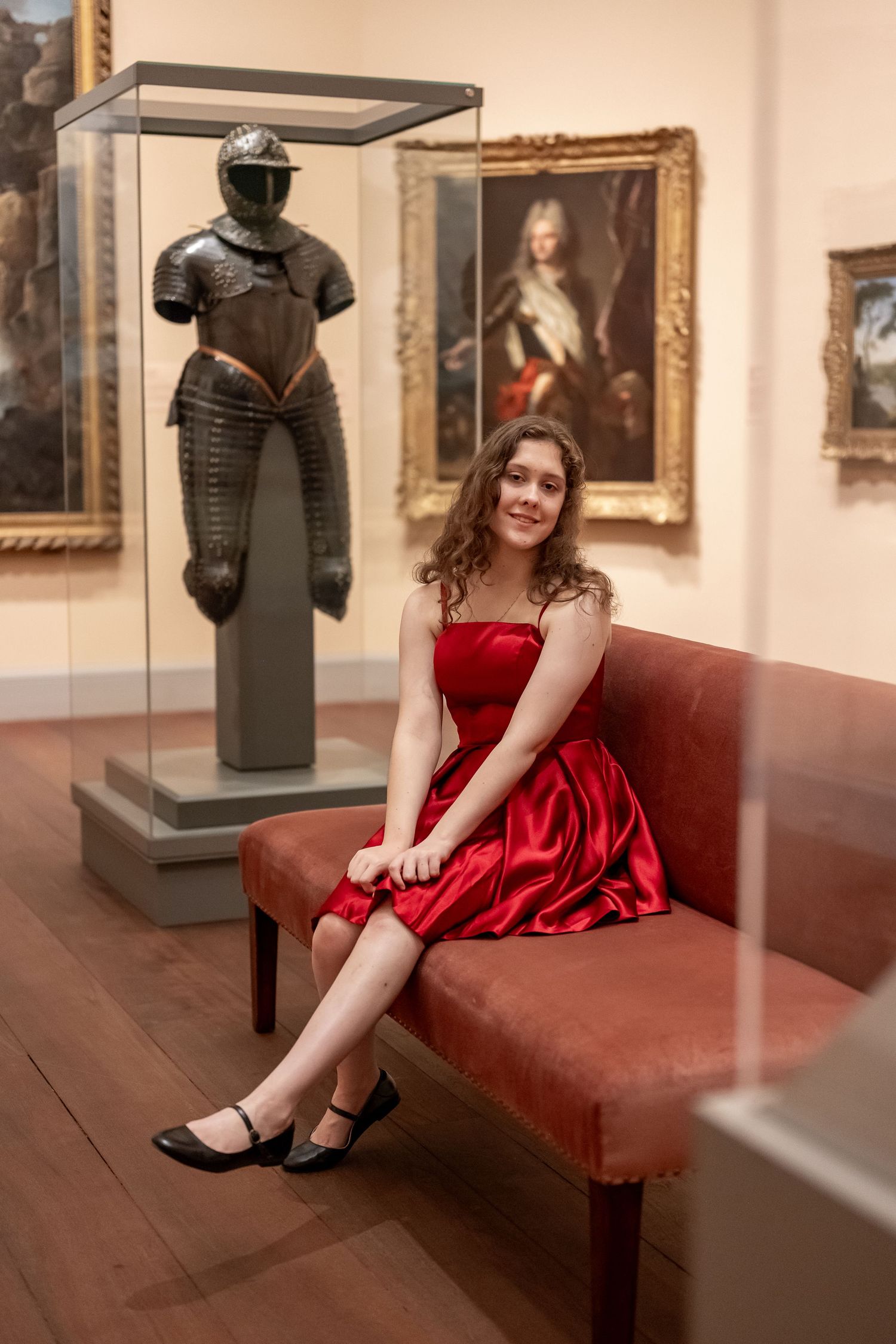 A person in a red dress poses elegantly on a vintage couch in an art gallery setting with sculptures and paintings visible.
