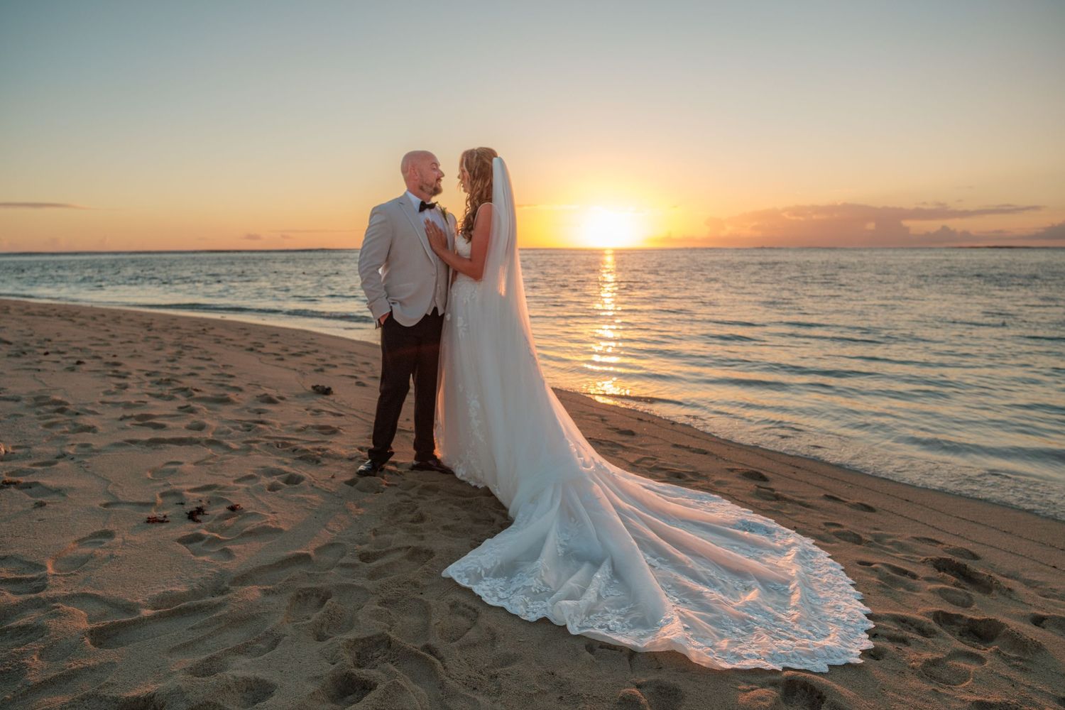 A romantic couple embraces on a tropical beach with palm trees and mountains in the background.