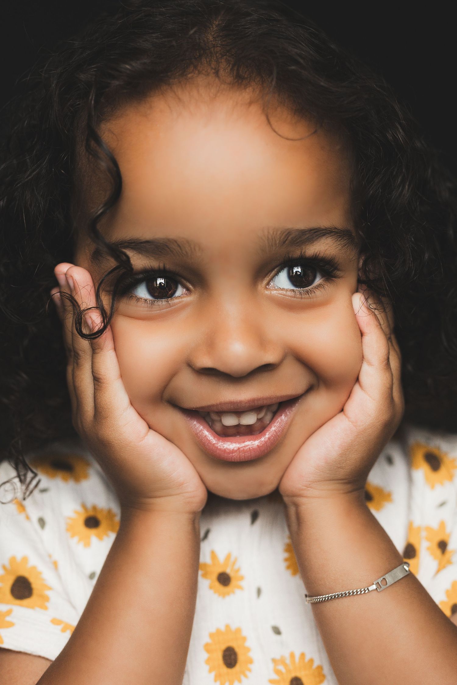 Happy young girl with curly hair cupping her face in her hands, wearing a white sunflower print dress.