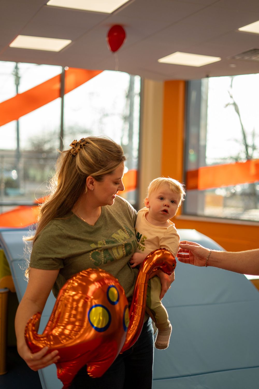 A woman holds a baby and a red balloon animal in a room with orange accents and fluorescent lighting.