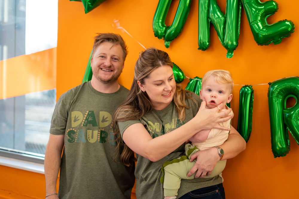 Family celebrates special moment against bright orange wall with green balloon decorations.