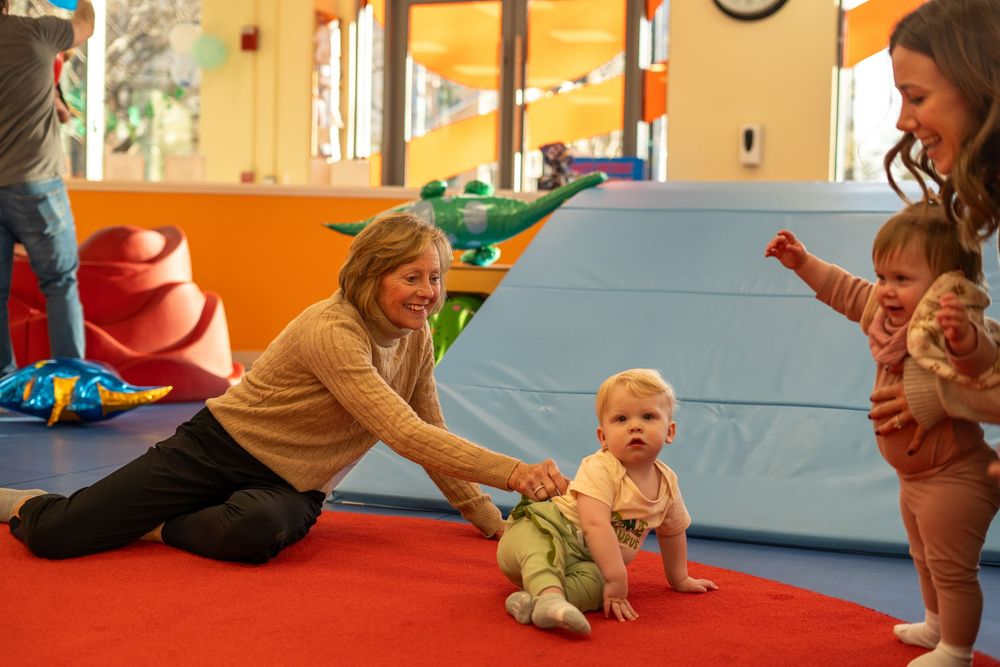 Babies and adults engage in floor activities at a bright orange-walled indoor play space.