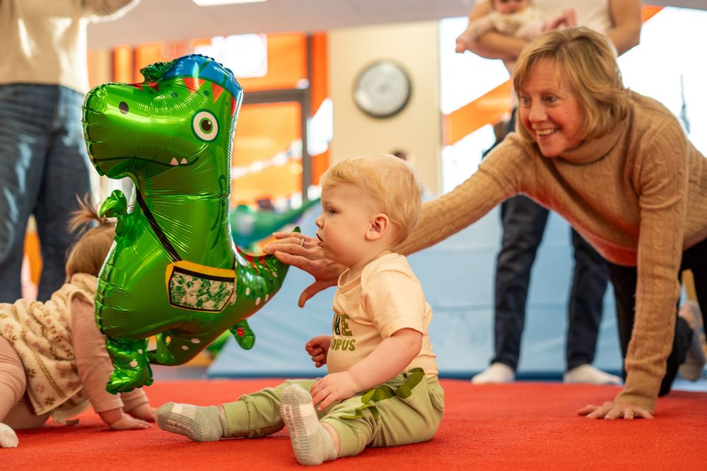 Baby in green outfit crawls on floor during playtime while adult supervises nearby.