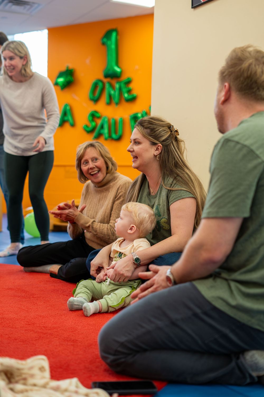 A group sits on a red carpet floor during an indoor activity session with colorful decorations on the wall behind them.