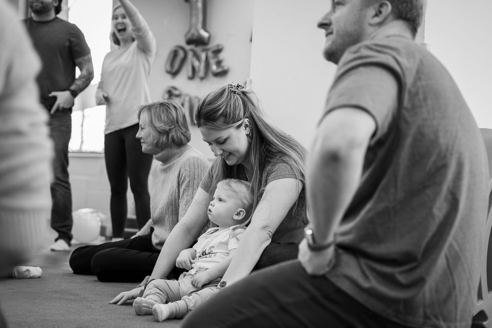 A toddler sits on someone's shoulders while watching a performance in a crowded indoor space.