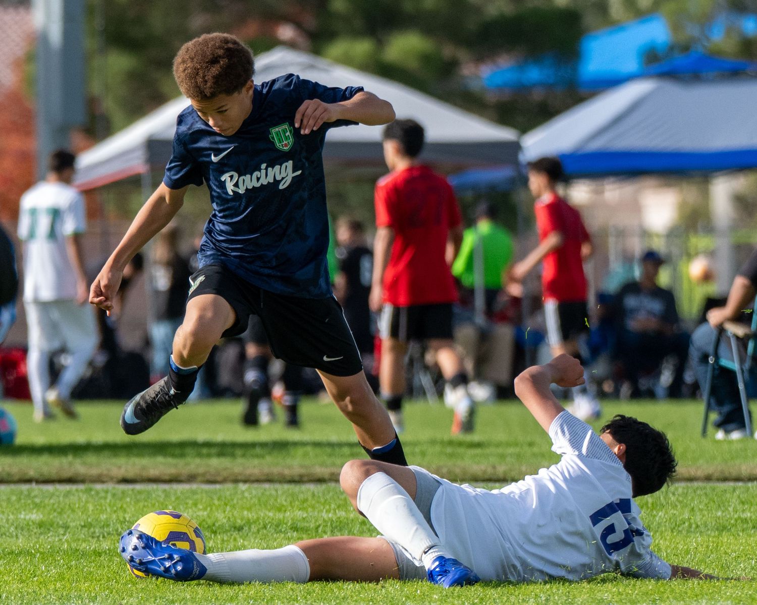 Youth soccer player attempts a shot while goalkeeper makes a sliding save on grass field.