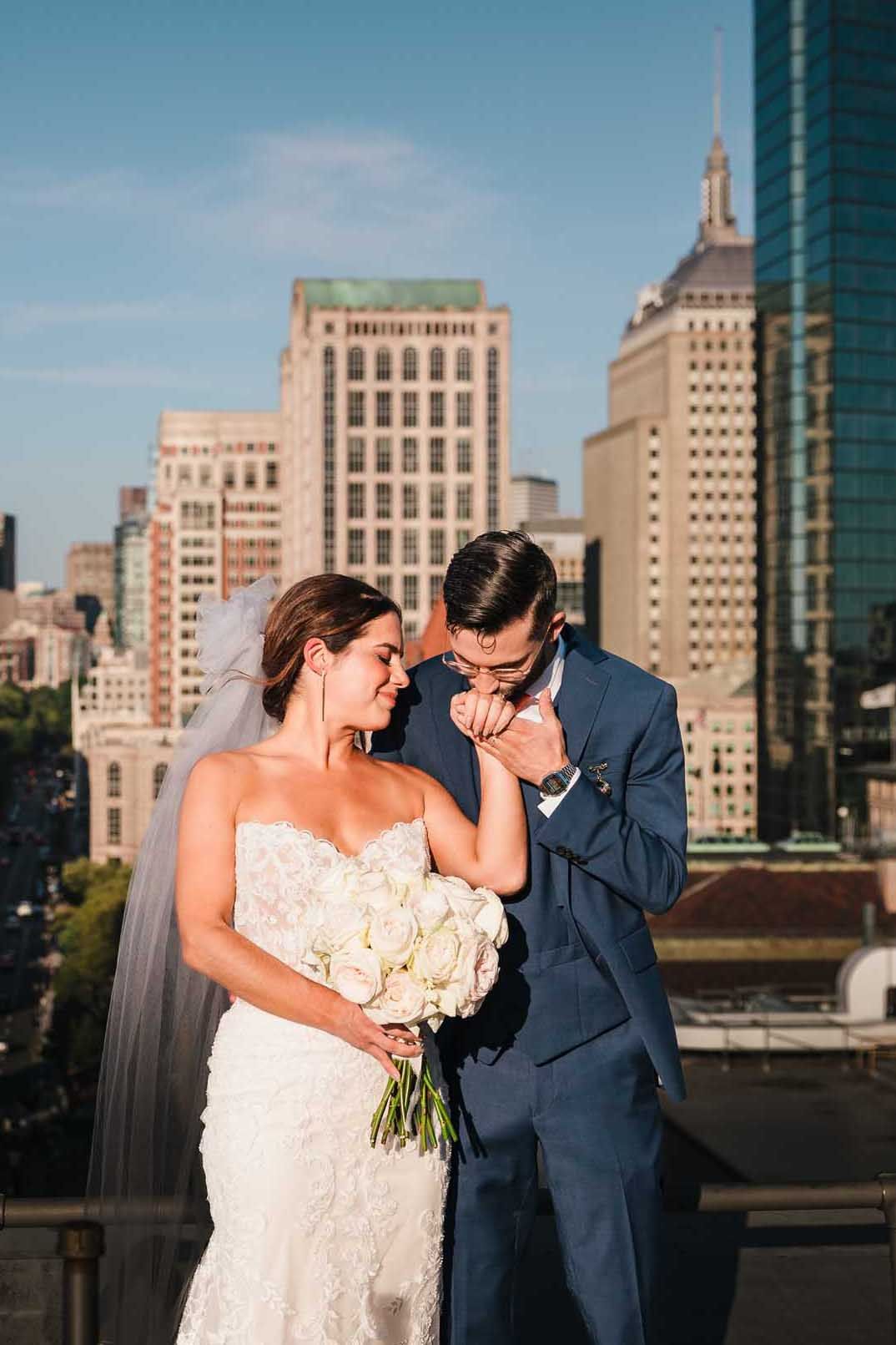 Groom kissing the bride’s hand during rooftop wedding portraits at the Lenox Hotel with the Boston skyline behind them