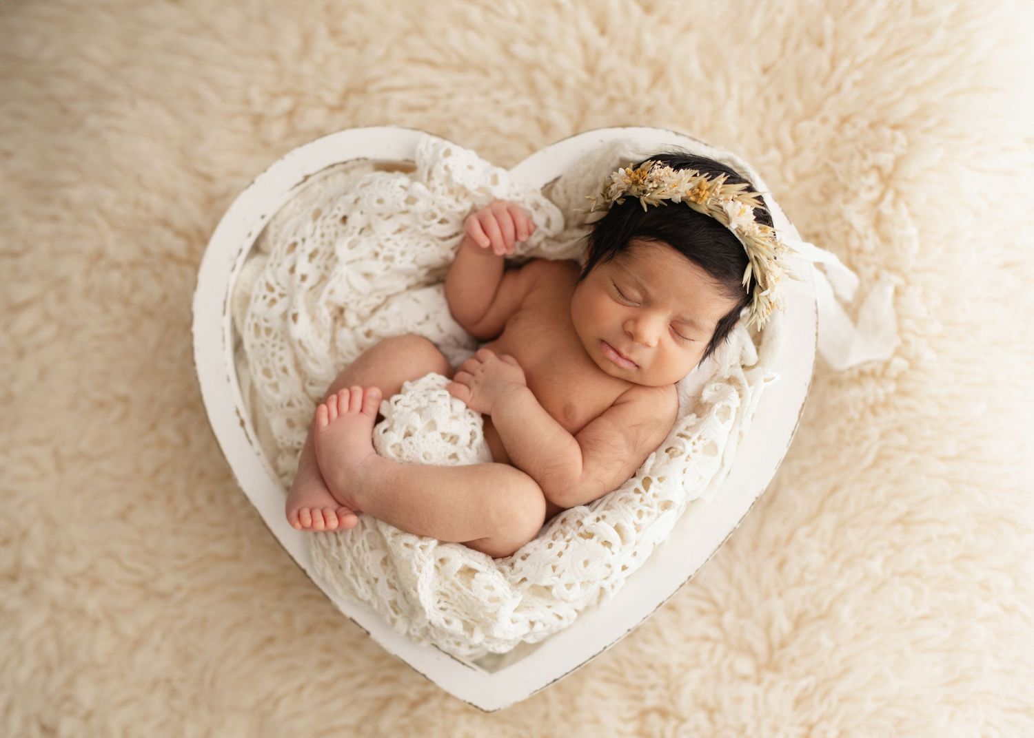 Sleeping newborn baby curled up in white heart-shaped basket on cream fur backdrop with lace headband. Elizabeth Lang Photography Surrey