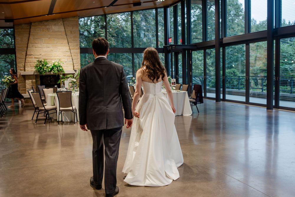 A couple in formal wedding attire walks through an elegant modern venue with floor-to-ceiling windows and natural light.