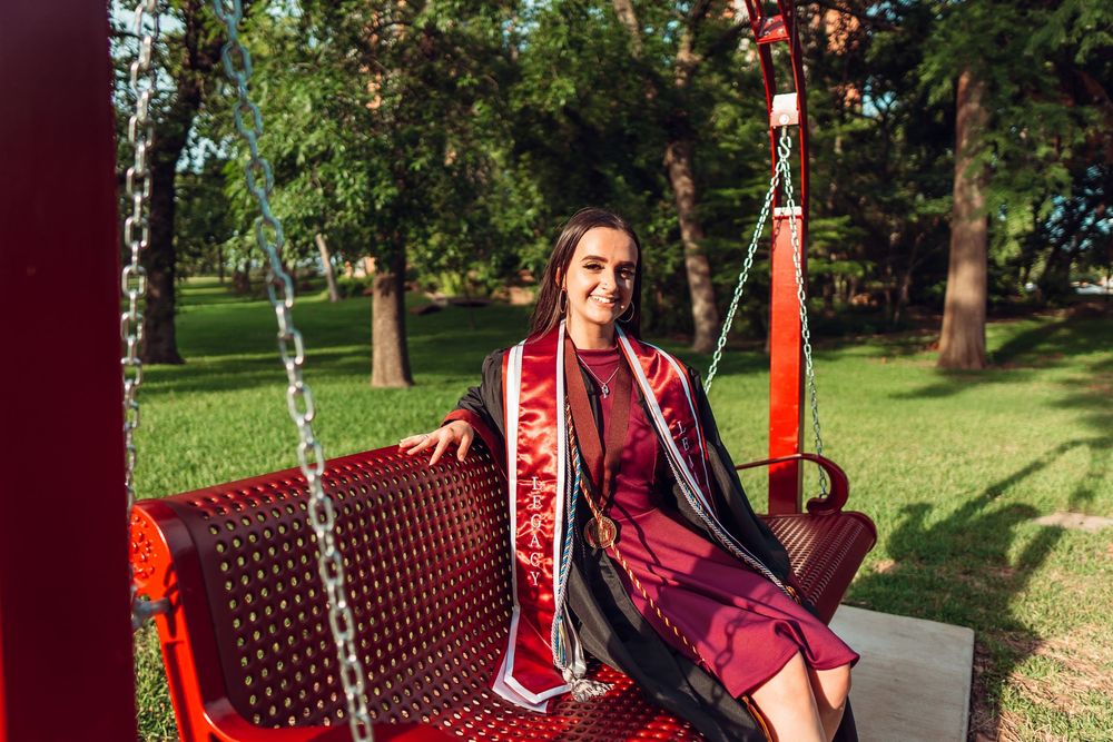 Graduate in formal attire poses on decorative red bench outdoors. TWU Graduate Portraits by Eric Aaberg Photography in DFW.