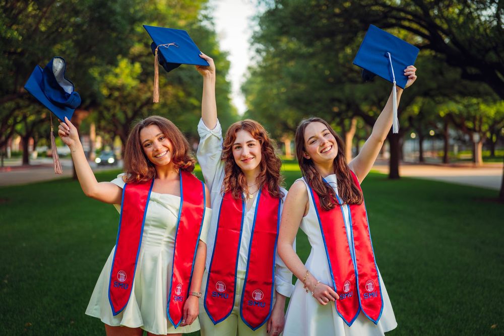 Three graduates in white dresses and red stoles celebrate by raising blue caps in tree-lined walkway. SMU Graduate Portraits by Eric Aaberg