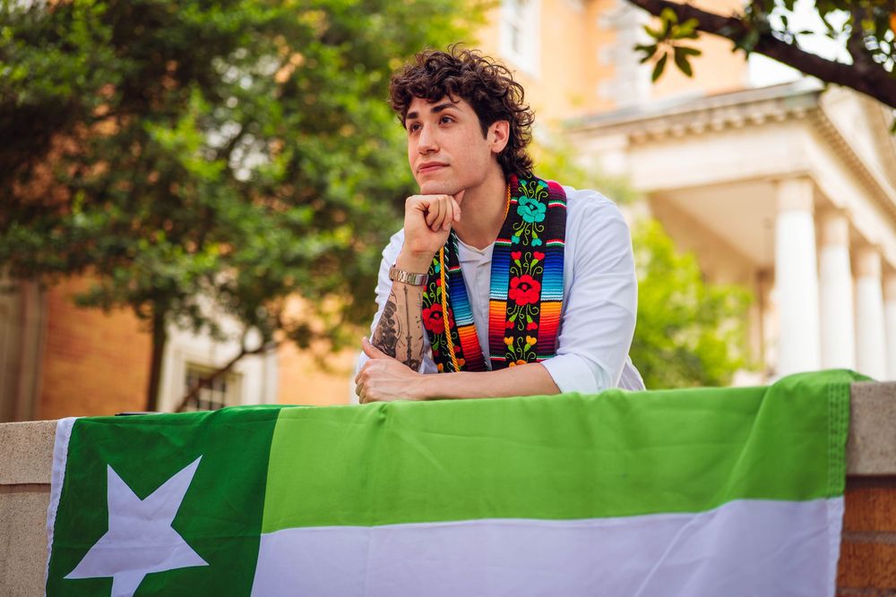 Student in colorful vest poses thoughtfully with green and white striped flag. UNT Graduate Portraits by Eric Aaberg Photography in DFW.