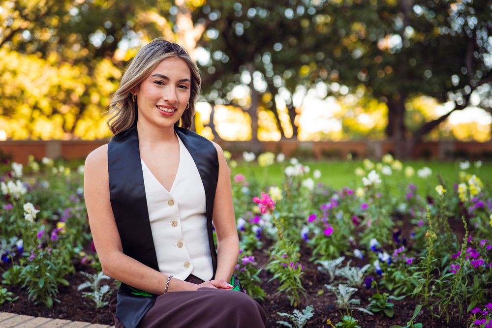 Graduate in formal attire sits among purple flowers during golden hour. UNT Graduate Portraits by Eric Aaberg Photography in DFW.