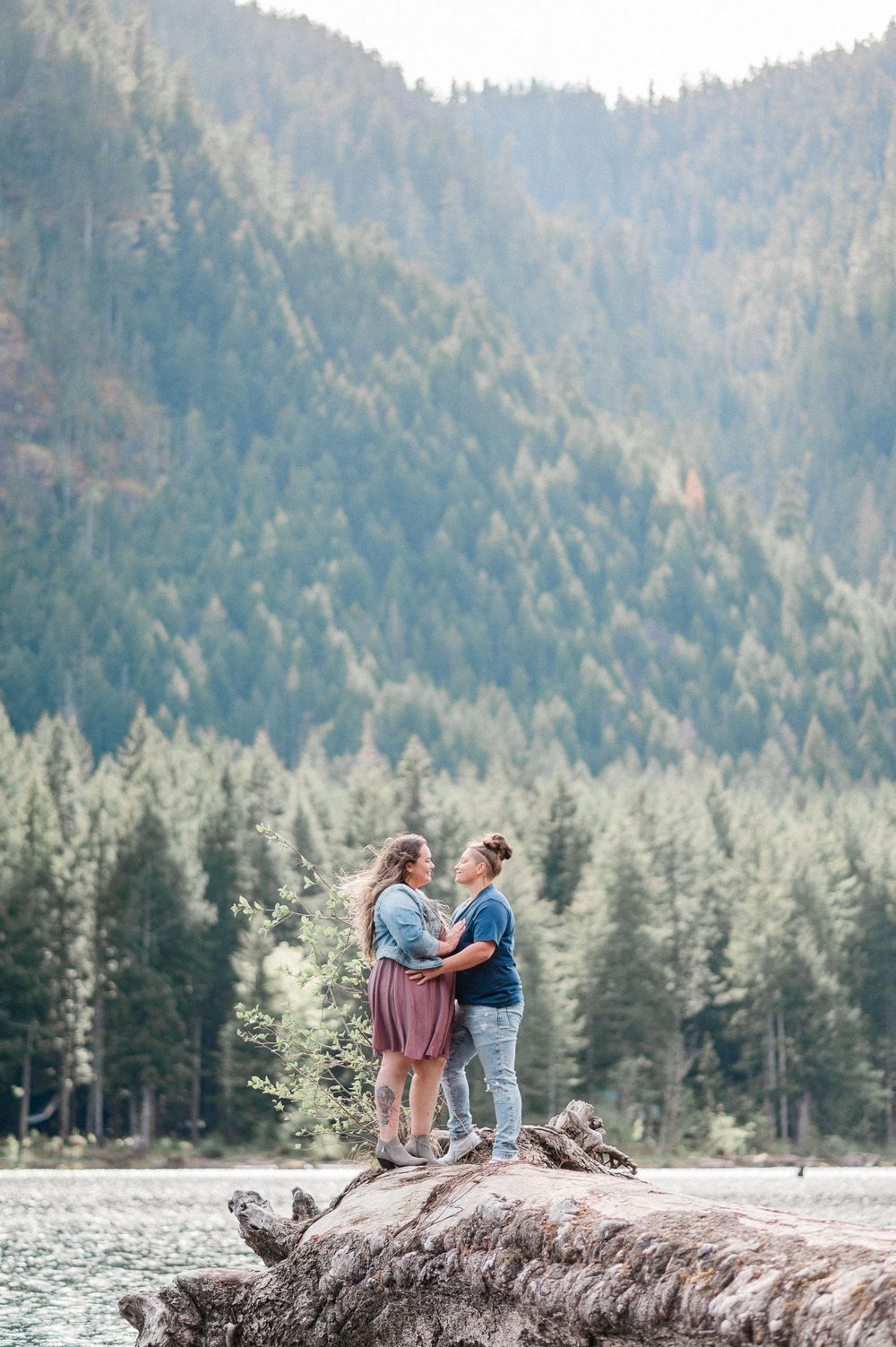 A couple stands on a fallen log by a serene mountain lake surrounded by evergreen forest on a sunny day
