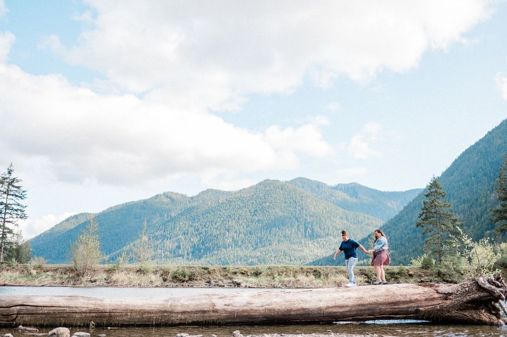Two people stand on a fallen log overlooking a scenic mountain landscape with evergreen trees and cloudy skies.
