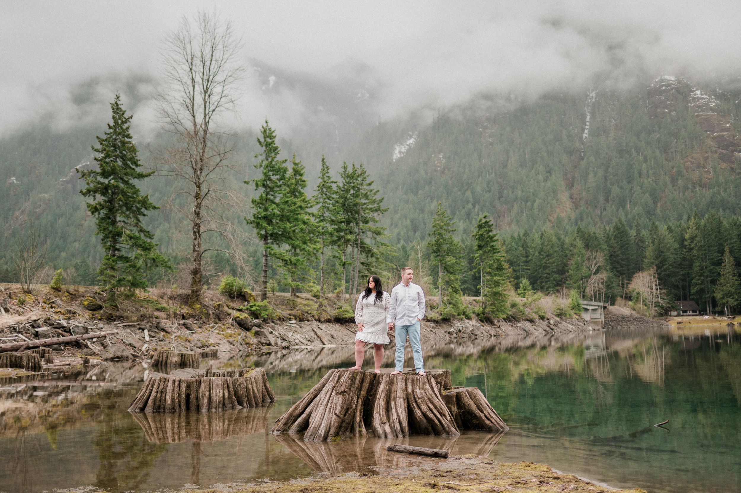 Couple in front of scenic mountain lake with tree stumps in foreground and evergreen forest along misty shoreline in Washington
