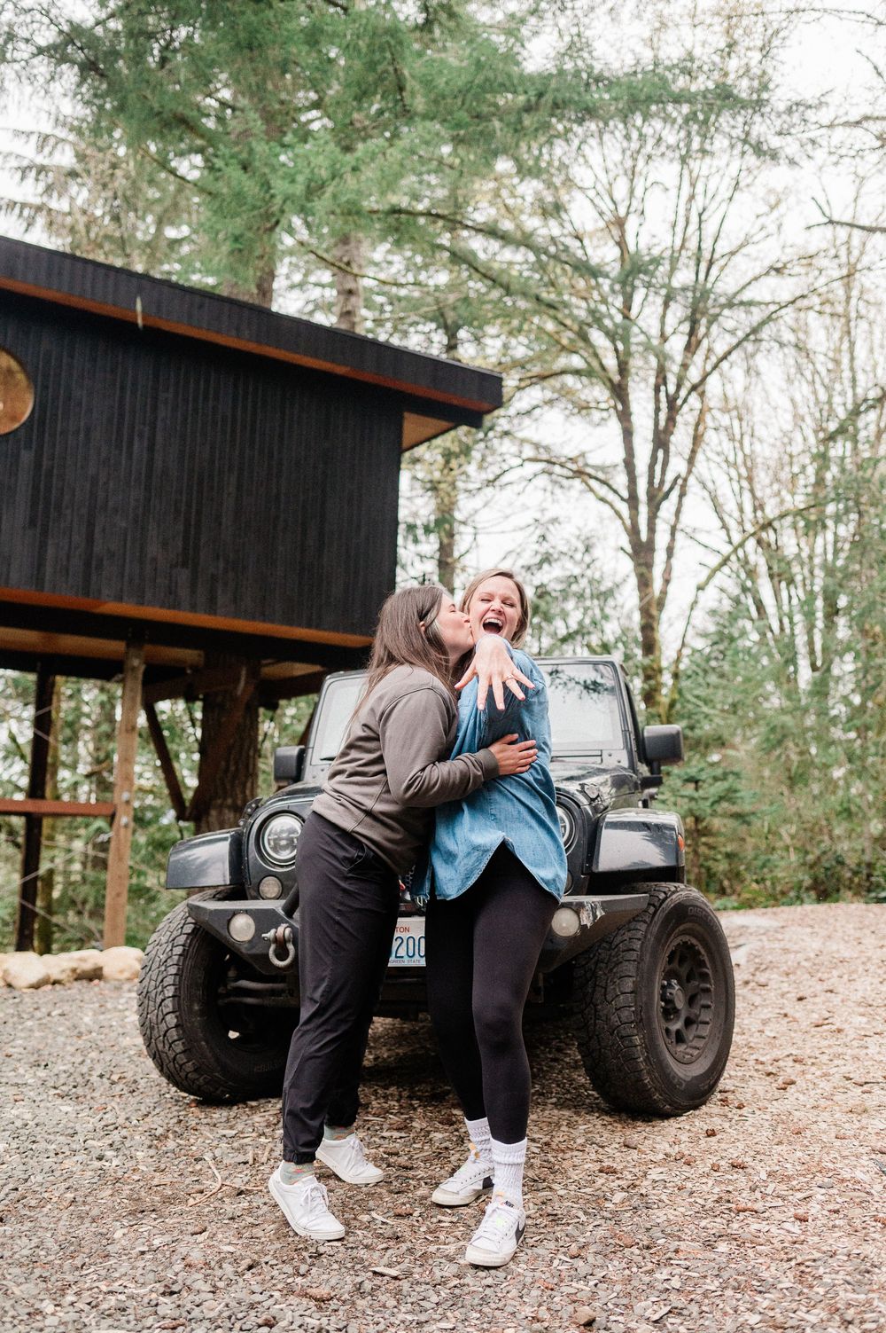 Two people share a joyful moment while leaning against a black Jeep parked near a rustic cabin in a wooded setting.