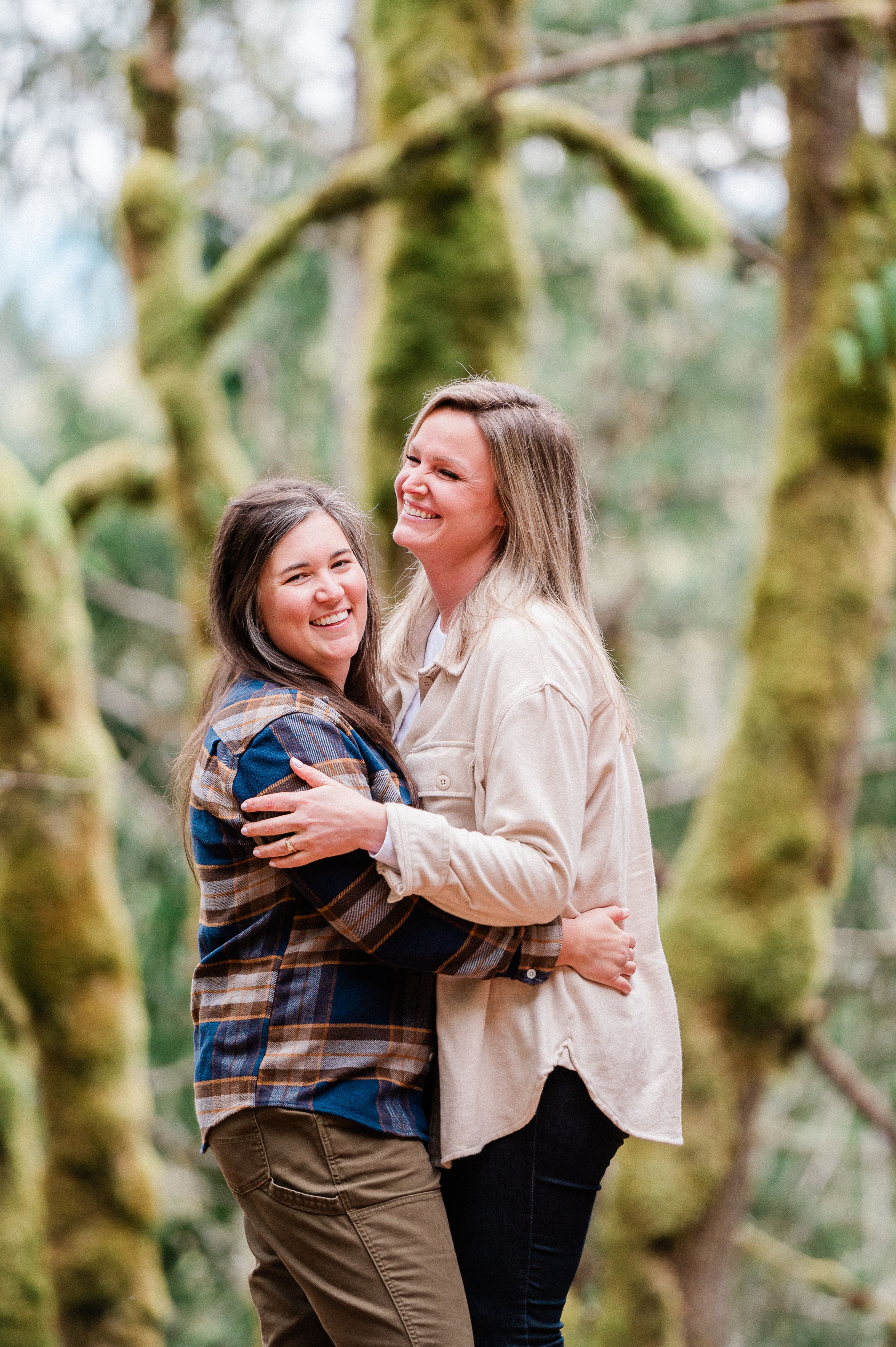 Two people share joyful embraces and laughter while standing among mossy trees in a forest setting.