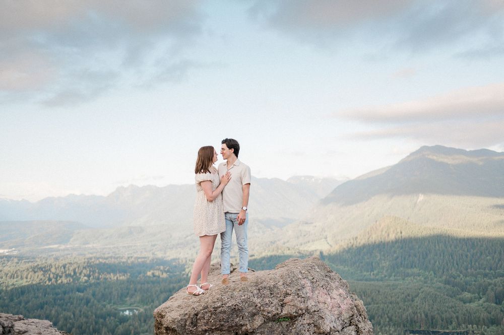 A couple stands together on a rocky mountain summit overlooking a scenic forest landscape at sunset.