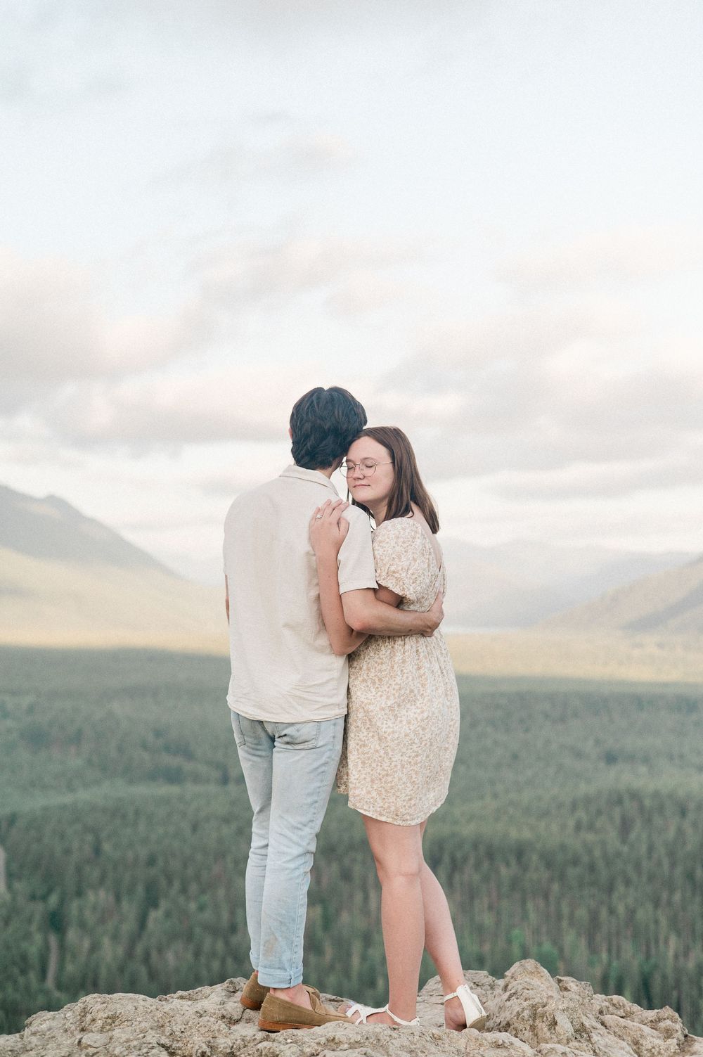 A couple shares an intimate moment while standing on a rocky overlook with a scenic mountain forest landscape at sunset.