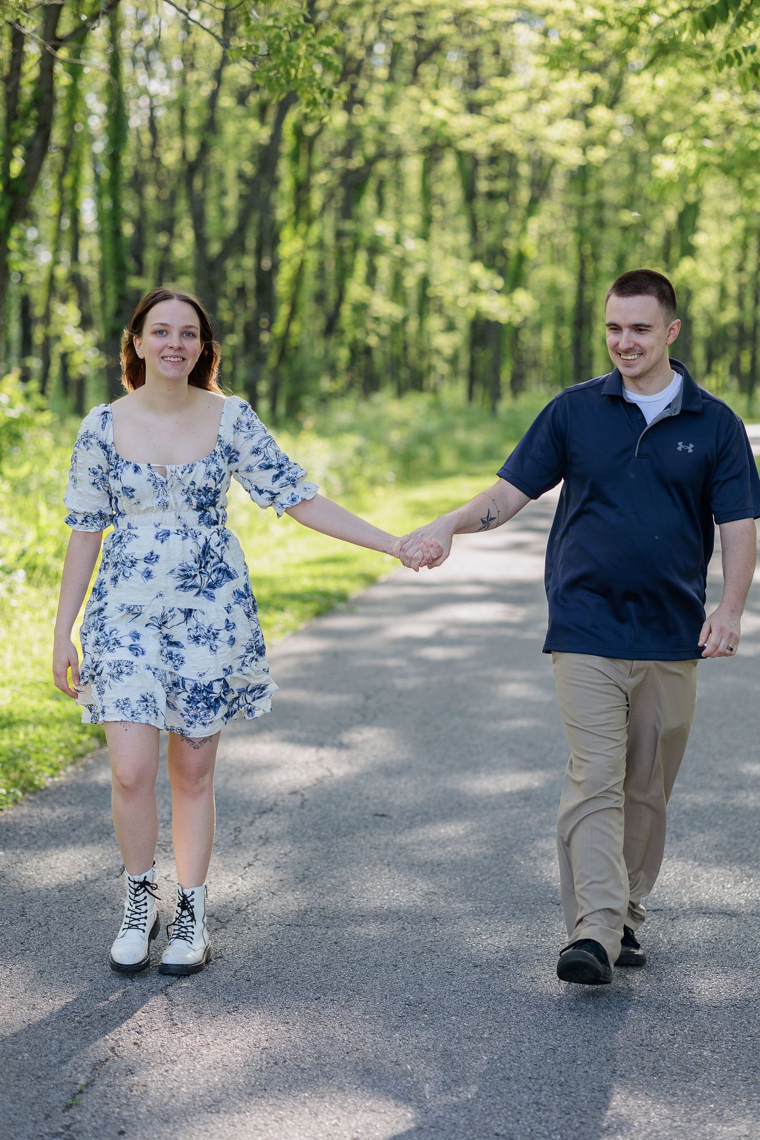 Two people in casual attire walk playfully on a tree-lined gravel path on a sunny spring day.