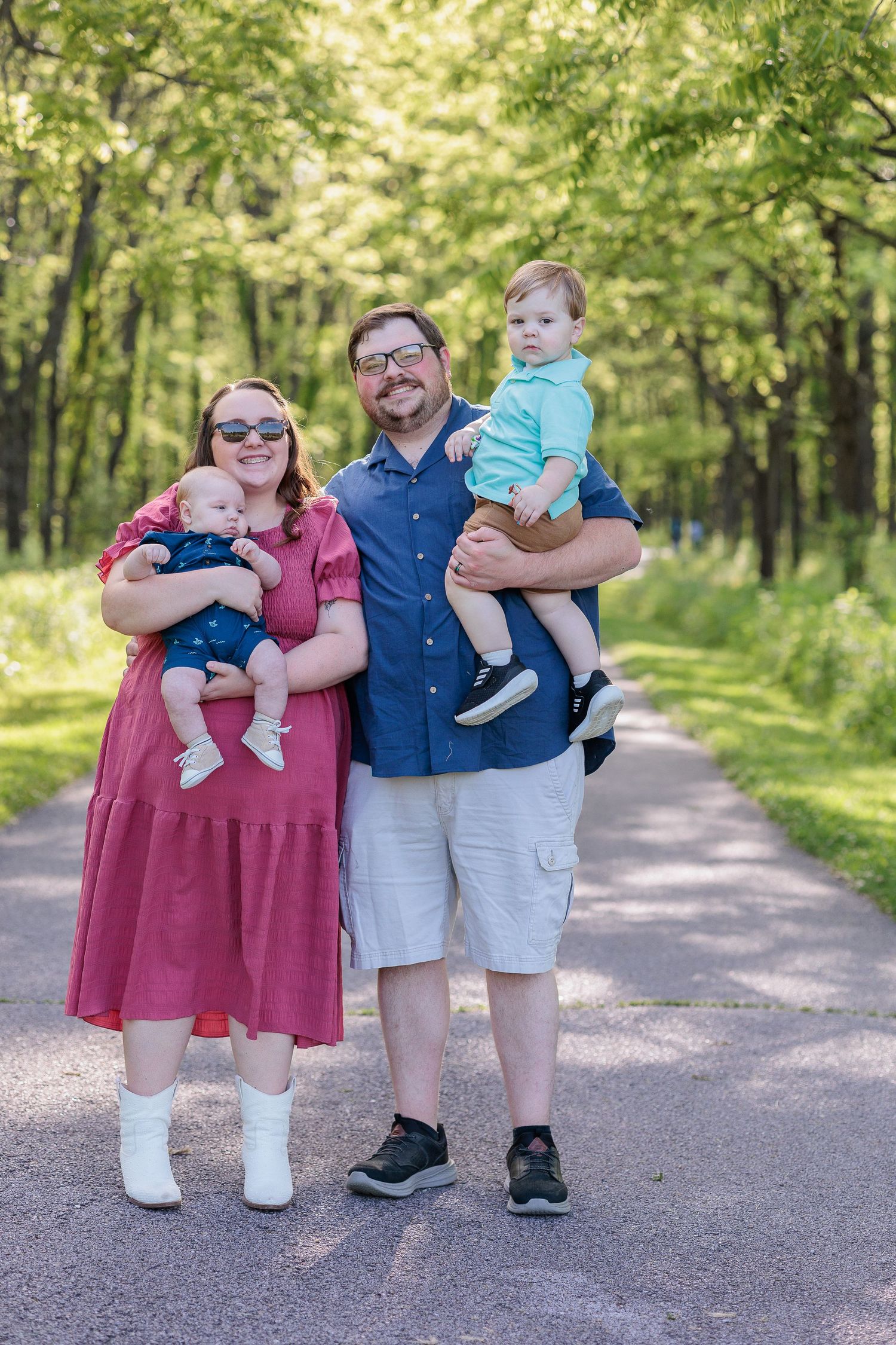 Family in coordinating blue and pink outfits poses together on a tree-lined path in a spring park setting.