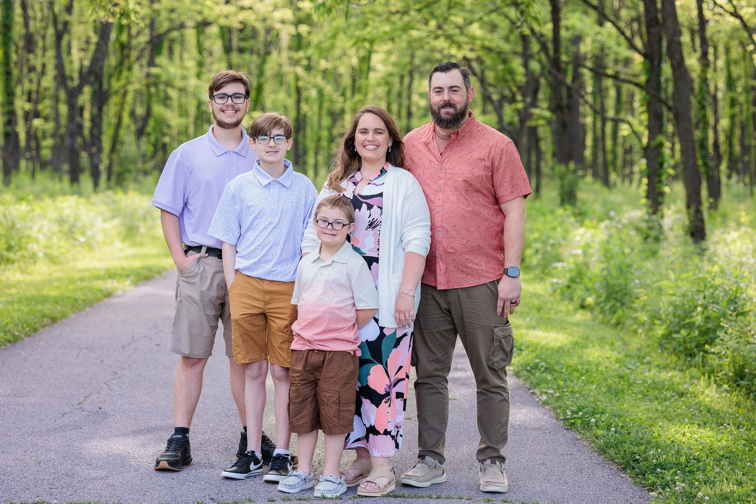 A family stands together on a tree-lined path in a lush green forest setting during spring photo session.