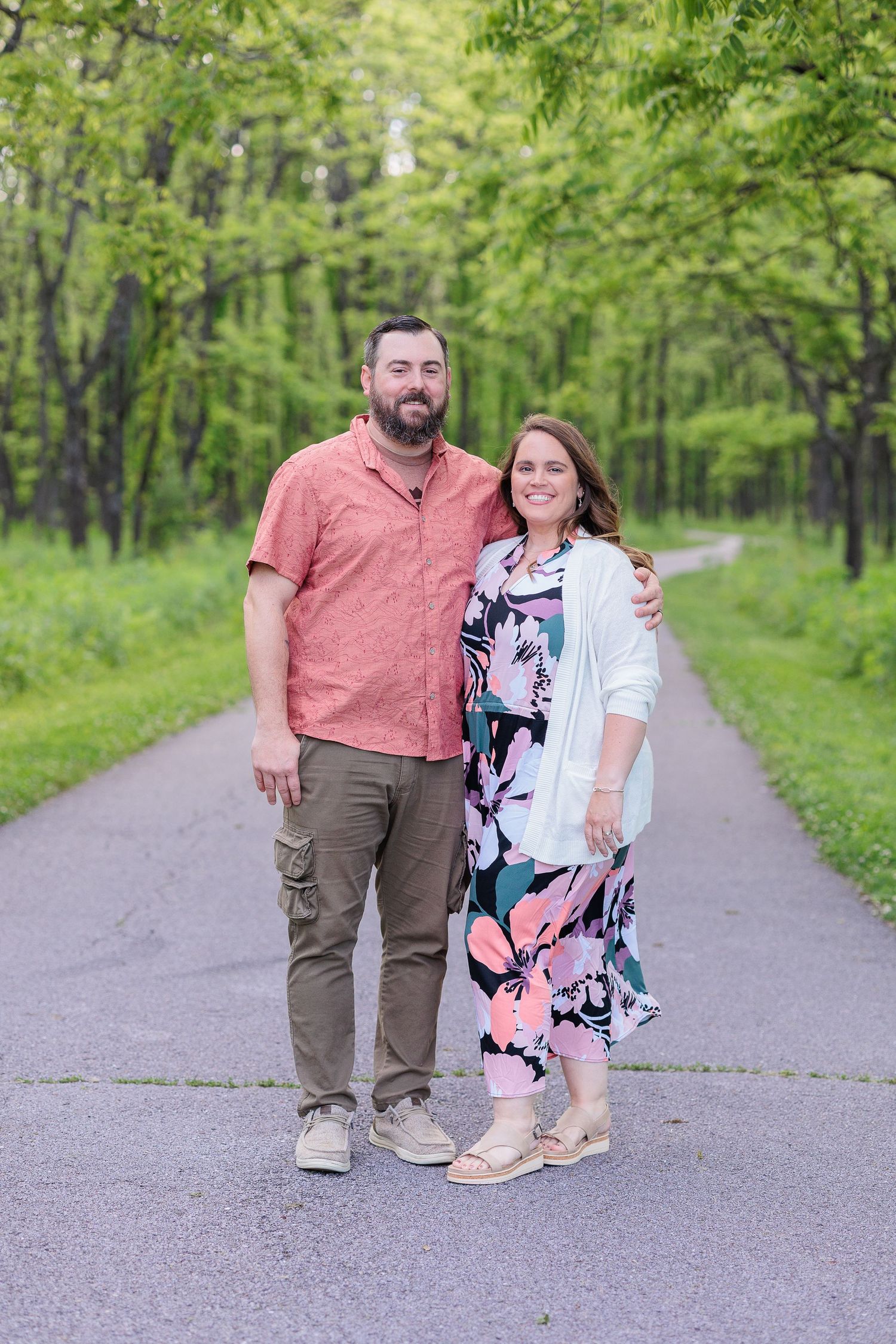 A couple stands together on a tree-lined path through a lush green forest on a spring day.