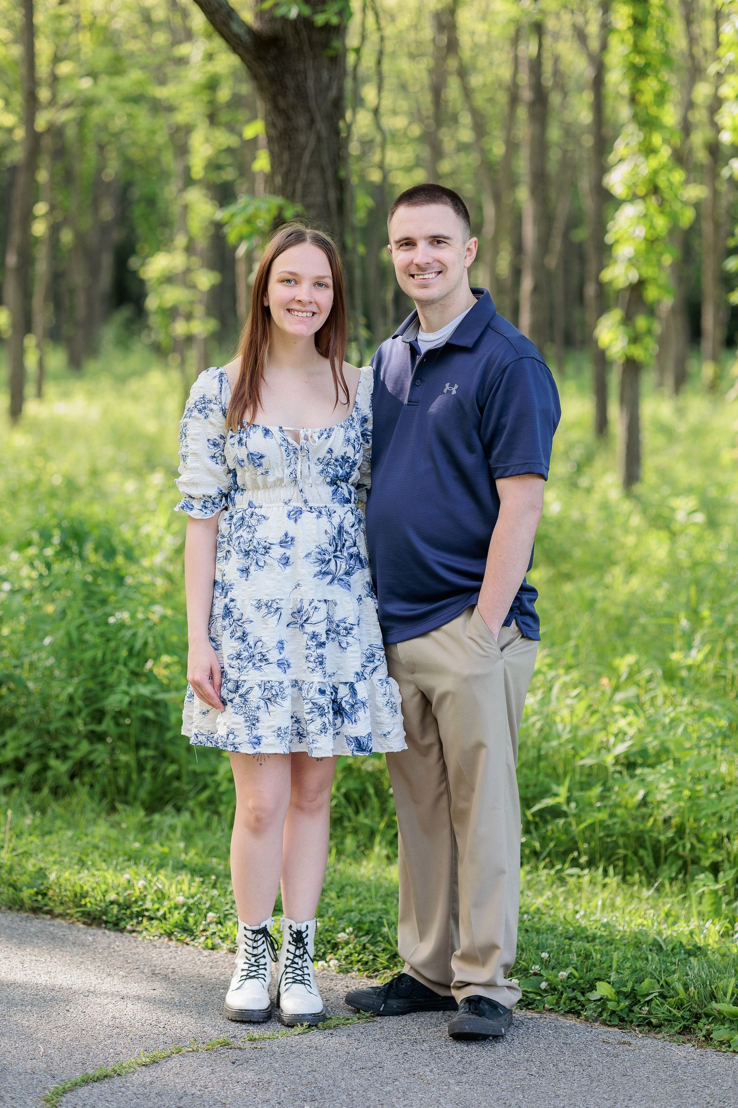 A couple stands together on a path in a lush green forest setting during a sunny spring day.