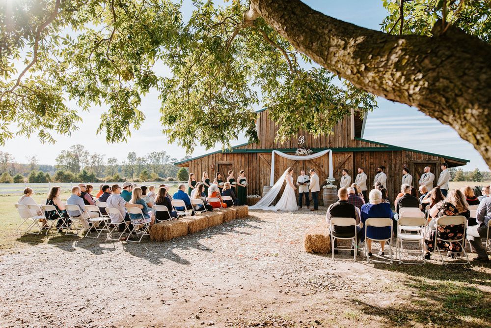 Wedding ceremony setup under large shady trees with white chairs and decorations in a rustic outdoor setting.