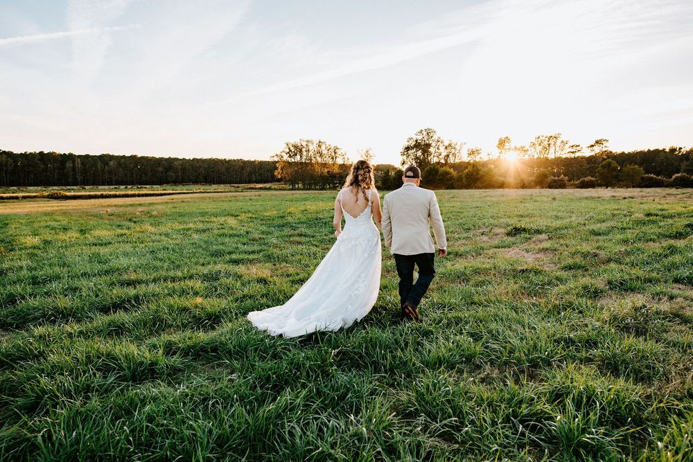 Romantic couple walks hand in hand through a golden field at sunset as her white wedding dress trails behind them.