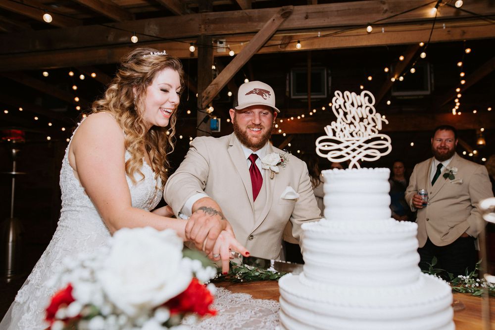 Couple cutting their elegant white wedding cake with chandelier lights twinkling in the background at reception venue.