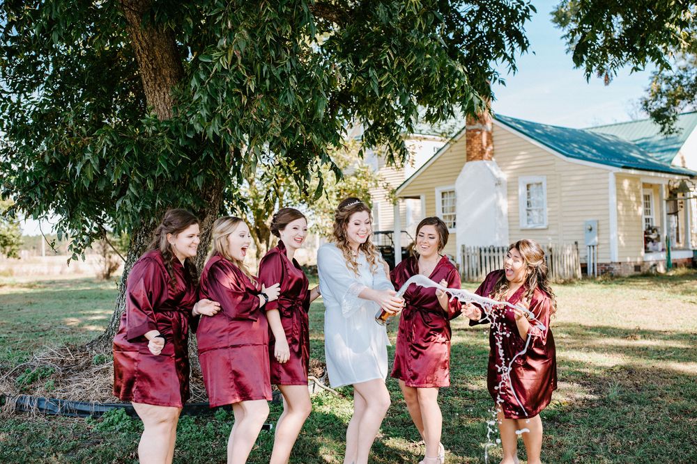 Wedding party in burgundy dresses and suits pose together outdoors on a sunny day near a white building.
