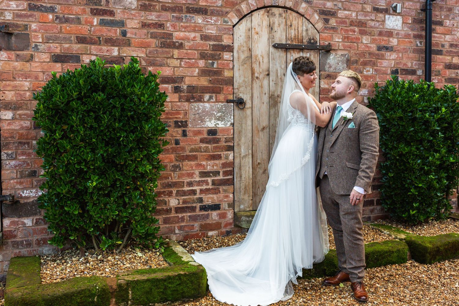 A romantic wedding couple shares a tender moment against a rustic brick wall backdrop with lush greenery.