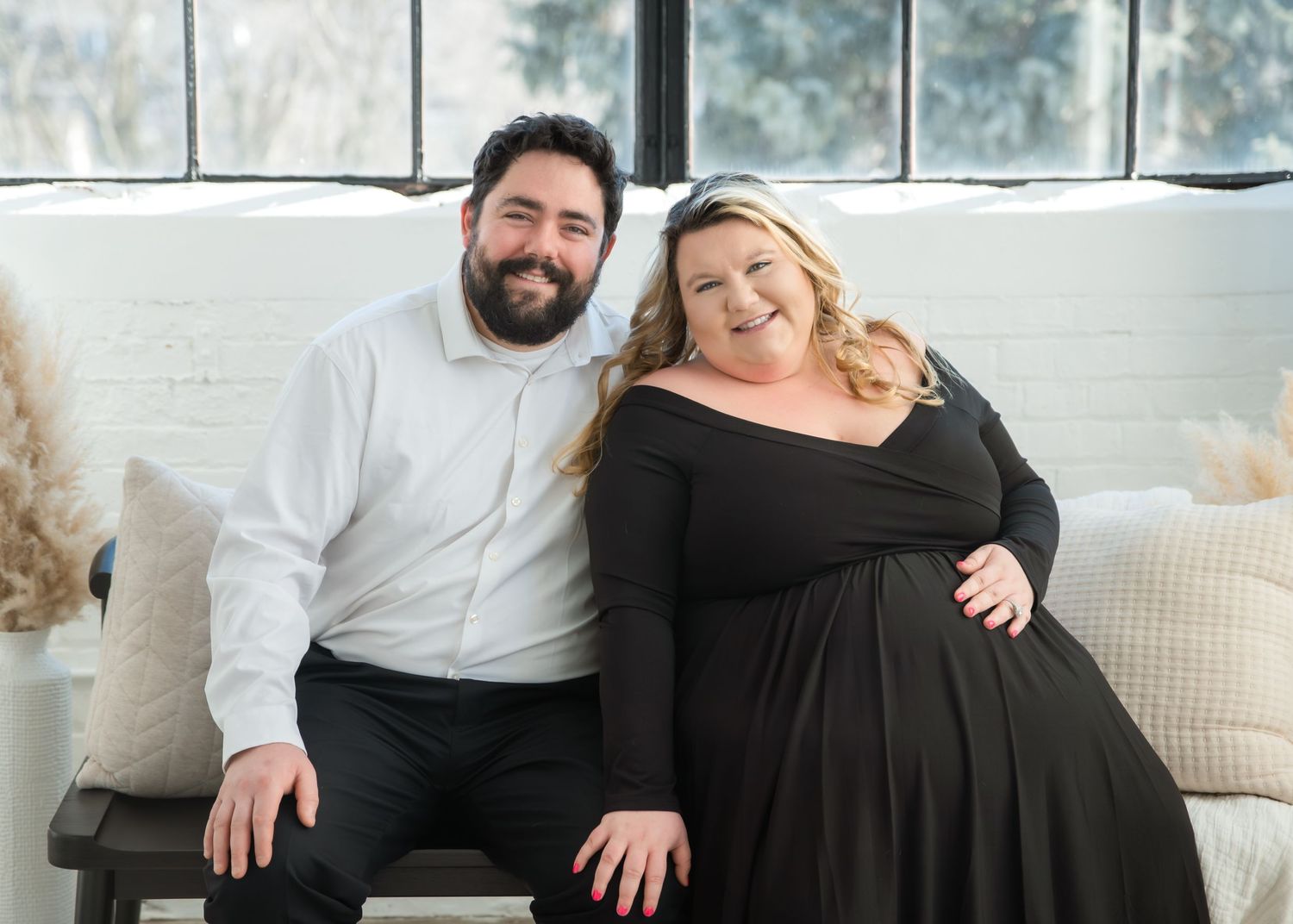 A couple sits together on a couch by a bright window, the woman wearing a black dress and the man in a white sweater.