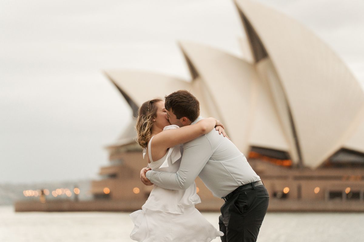 A couple shares a romantic hug together with the Sydney Opera House visible in the background during a proposal photoshoot