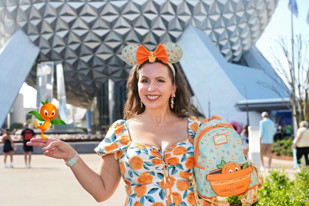 Woman in orange and white floral dress poses with a drink at an outdoor event.