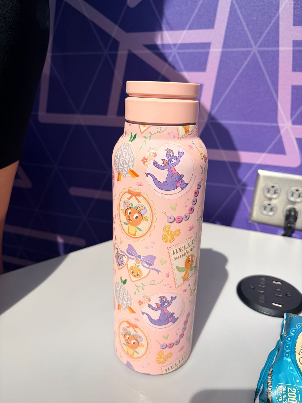 A pink ceramic mug with whimsical ghost and pumpkin Halloween designs sits on a counter near an electrical outlet.