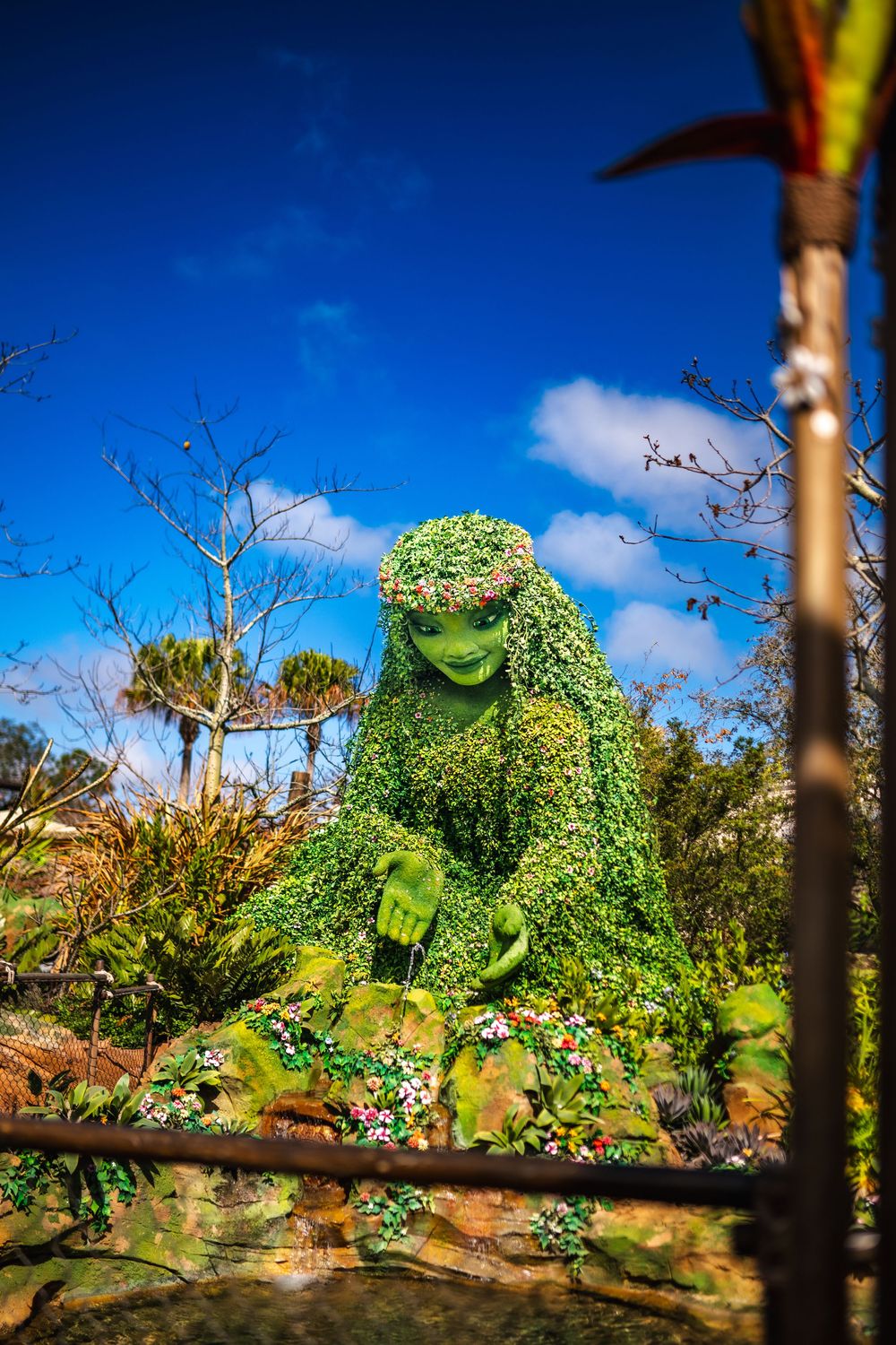 Green vines grow along a rustic wooden fence against a bright blue sky with white clouds on a sunny day.