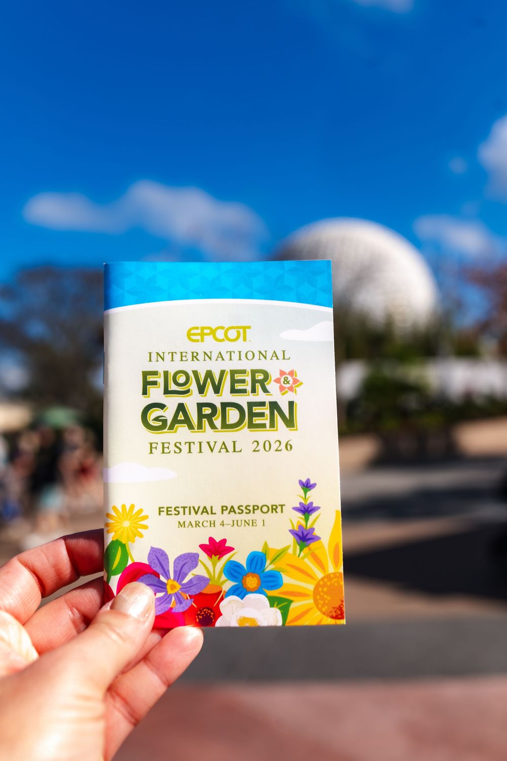 Hand holding Epcot International Flower & Garden Festival 2026 passport with Spaceship Earth visible in background.