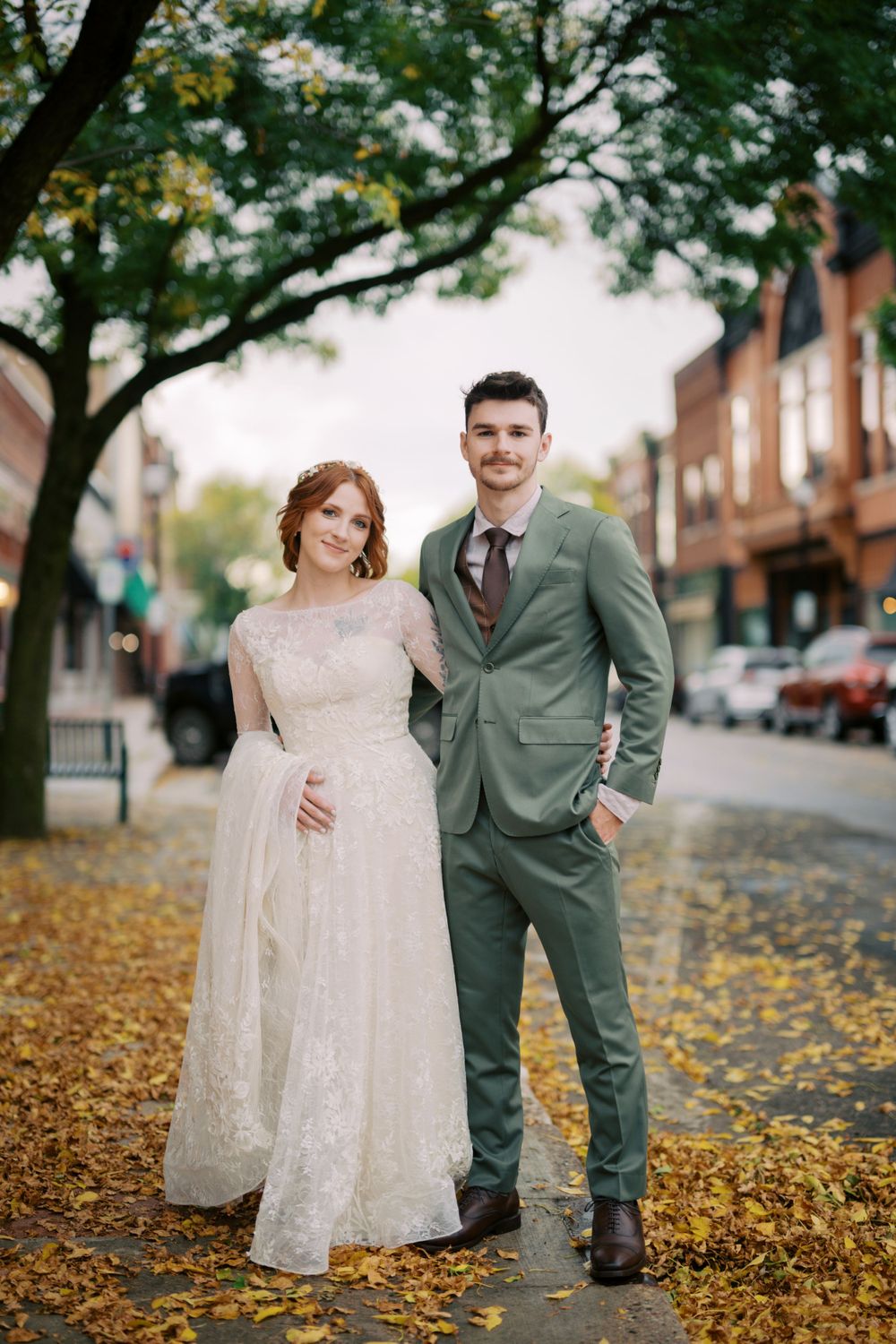 A couple poses on a fall day with autumn leaves on a city street lined with brick buildings and trees.