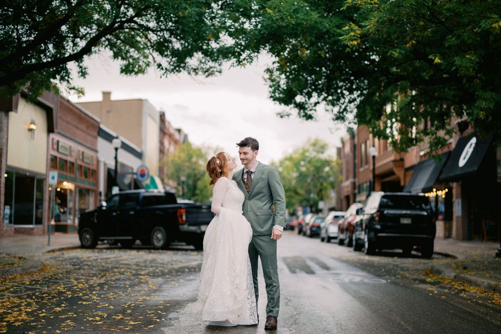 A couple poses together on a tree-lined street with autumn leaves scattered on the pavement during their wedding day.