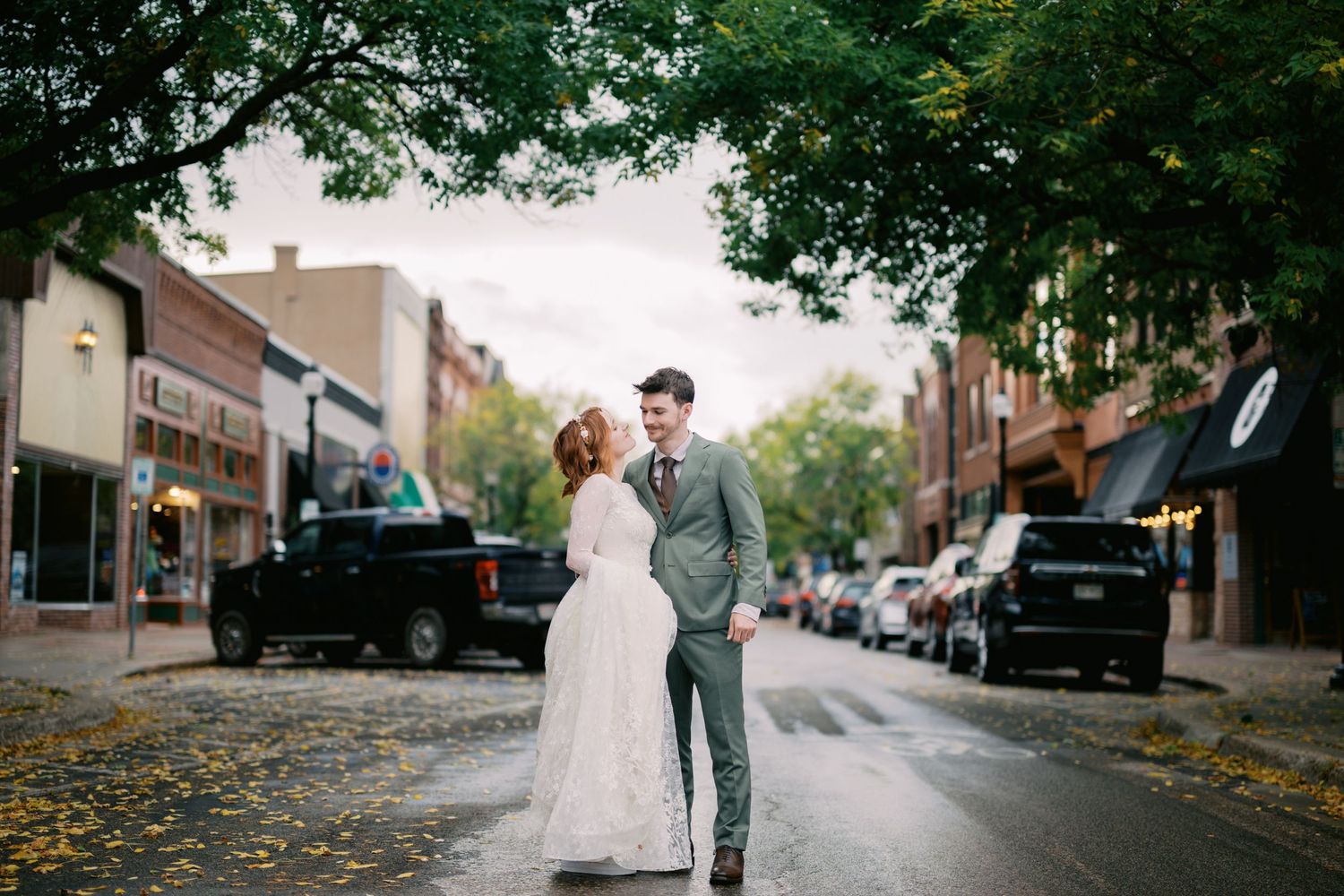 A couple poses together on a tree-lined street with autumn leaves scattered on the pavement during their wedding day.