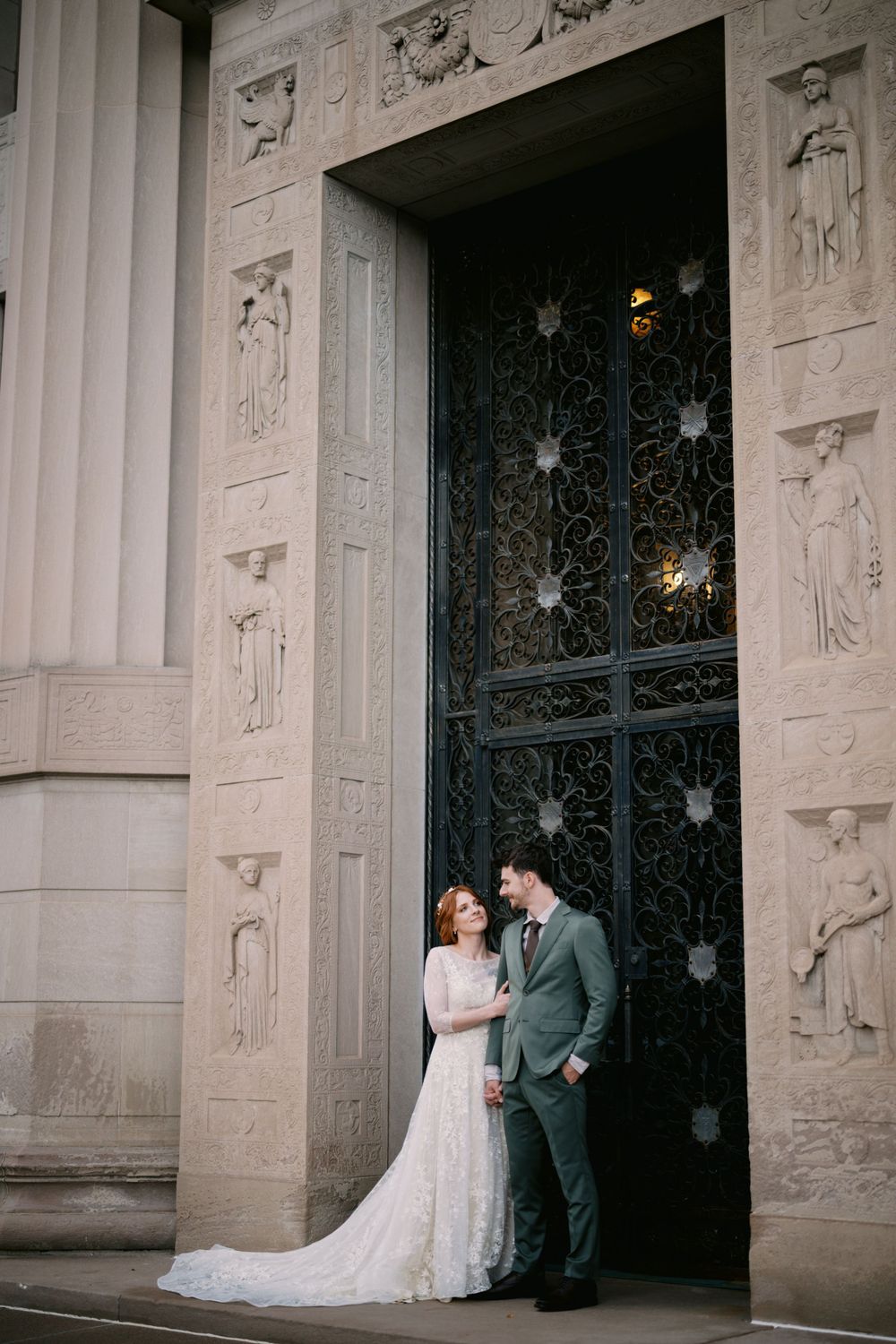 A couple stands in elegant wedding attire against ornate cathedral doors with decorative stone carvings and metalwork.