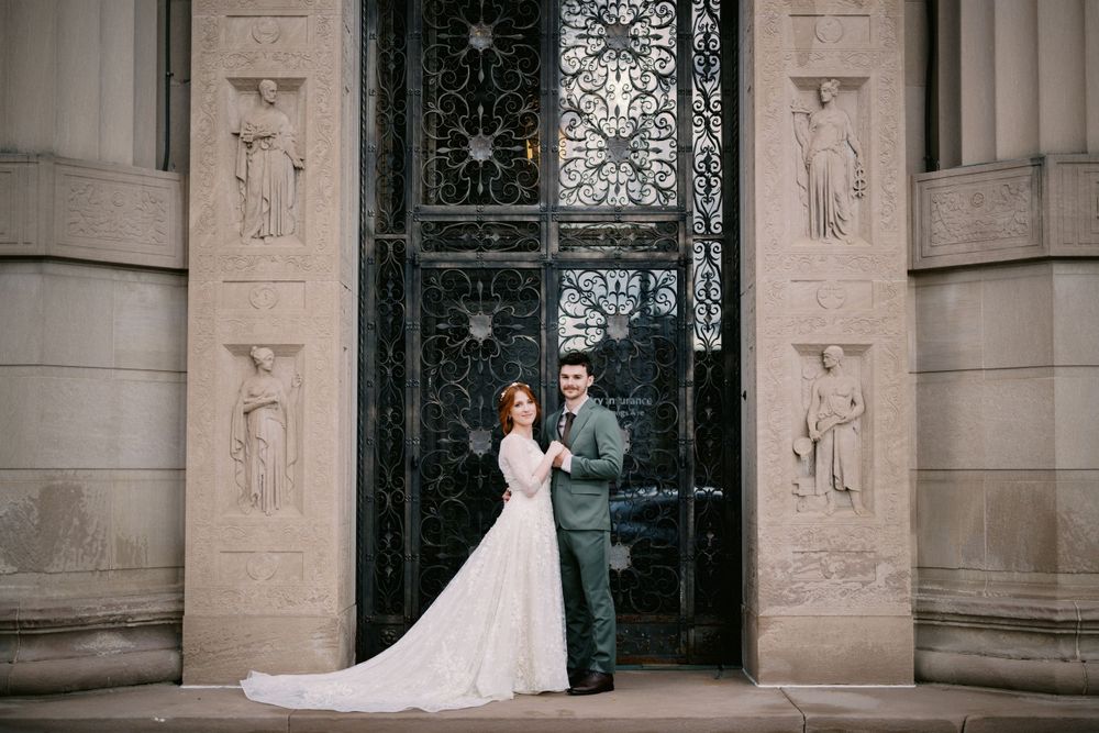 A couple in wedding attire stands before an ornate stone doorway with decorative carved figures on the columns.