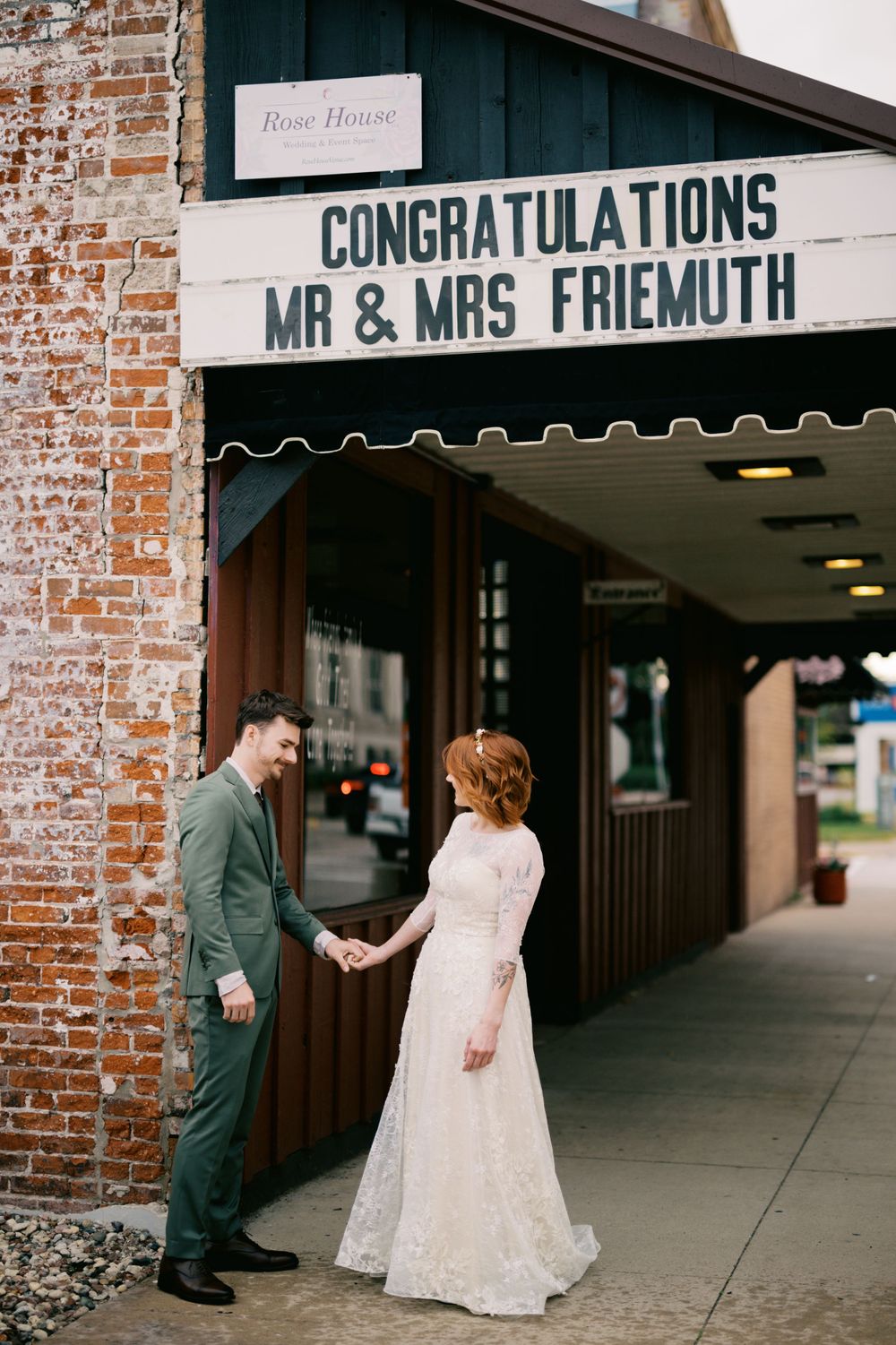 A couple stands under a congratulatory marquee sign for Mr & Mrs Friemuth outside a brick building.