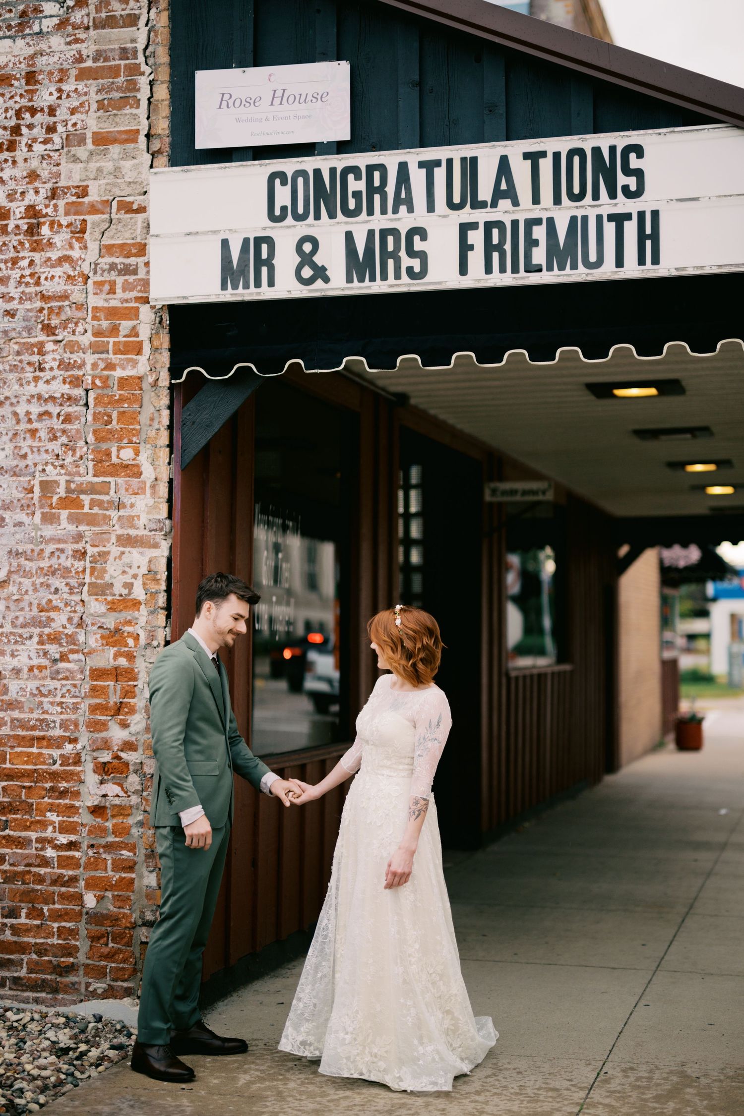 A couple stands under a congratulatory marquee sign for Mr & Mrs Friemuth outside a brick building.