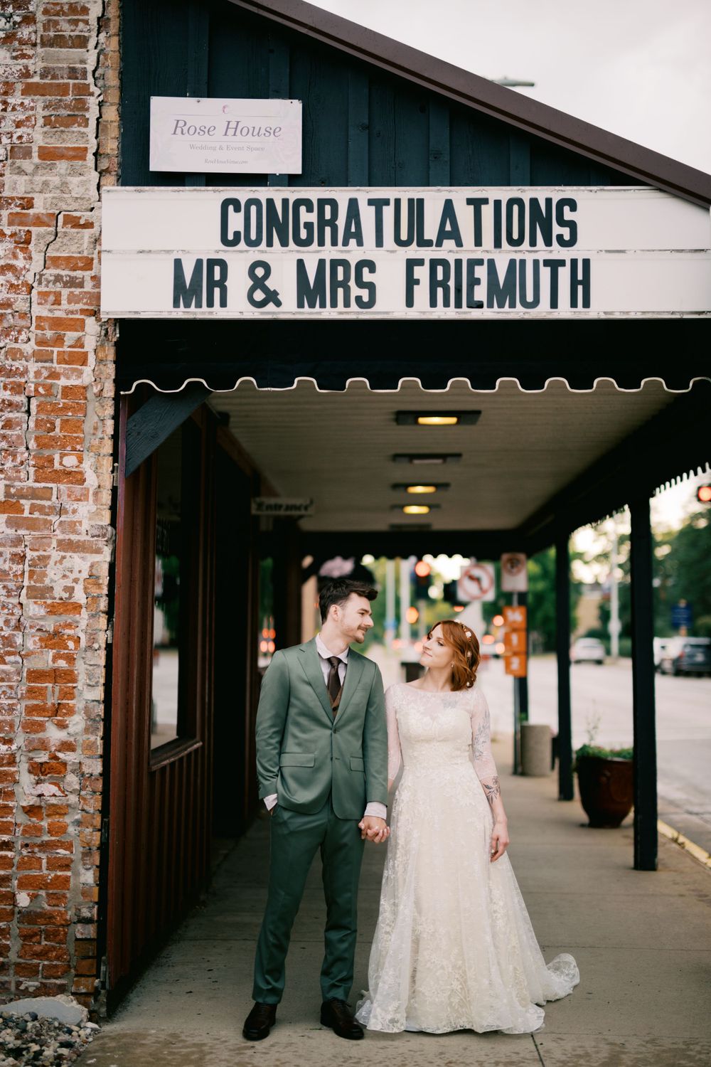 Wedding couple stands beneath a congratulatory sign with their name outside a brick building with an awning.