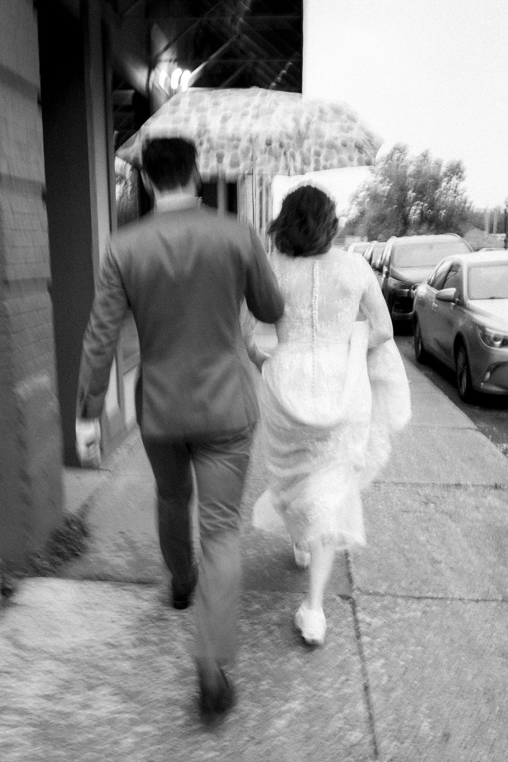 A black and white photograph of a couple walking down a sidewalk, with the person in white wearing formal attire.